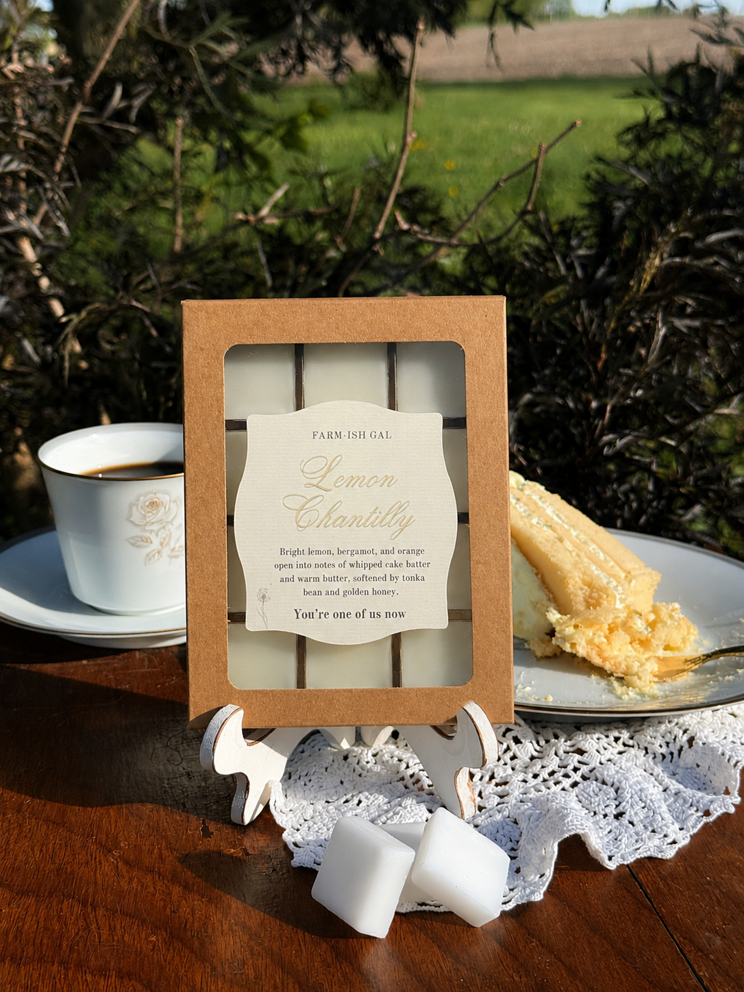 wax melts in a Kraft box with window and tag saying Lemon Chantilly. Cup of coffee, piece of cake and 3 square wax melts on wooden table set around wax melt box.
