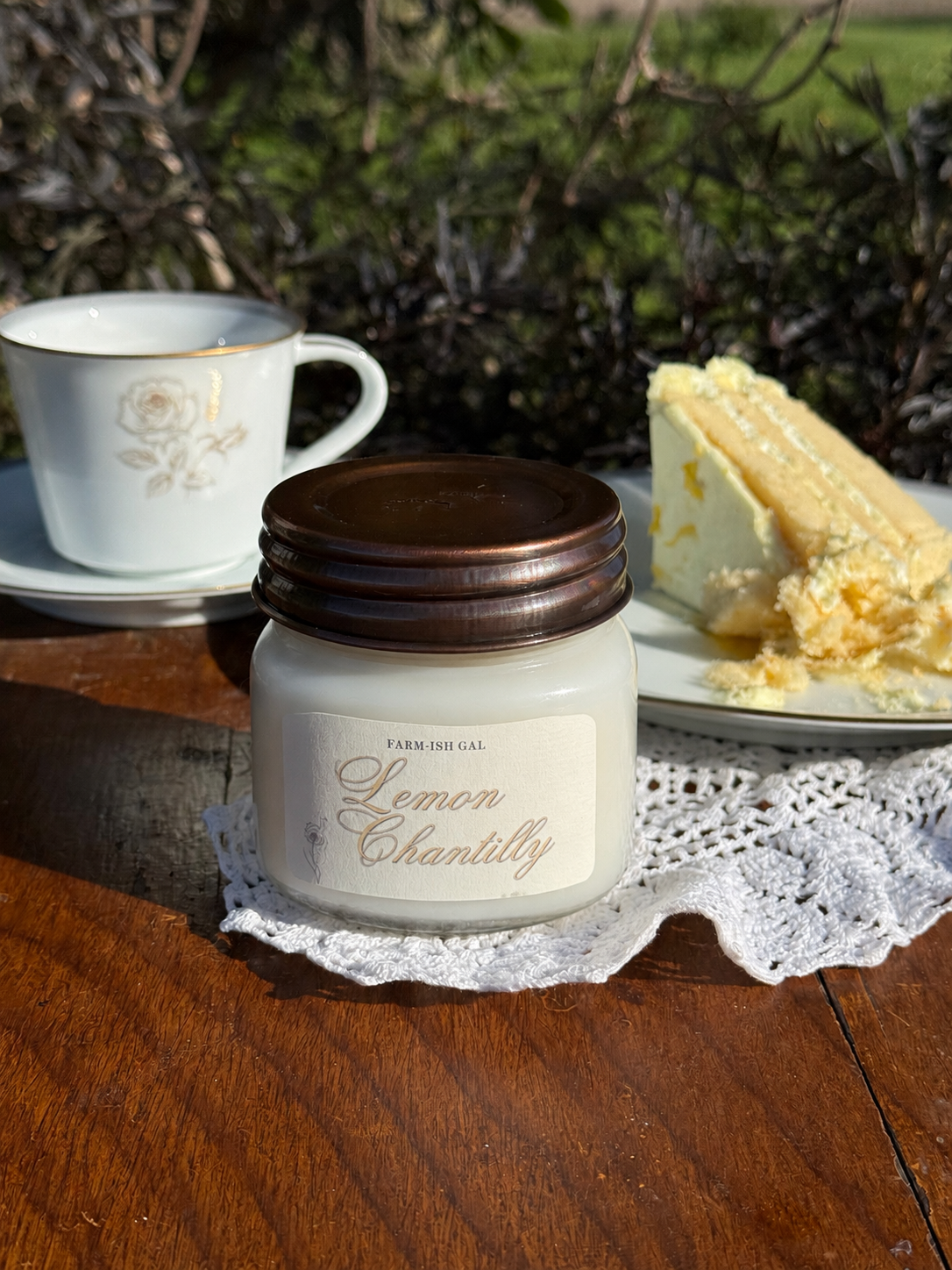 Candle with a label that says 'Lemon Chantilly' with a cup of coffee and piece of cake on a wooden table outdoors.
