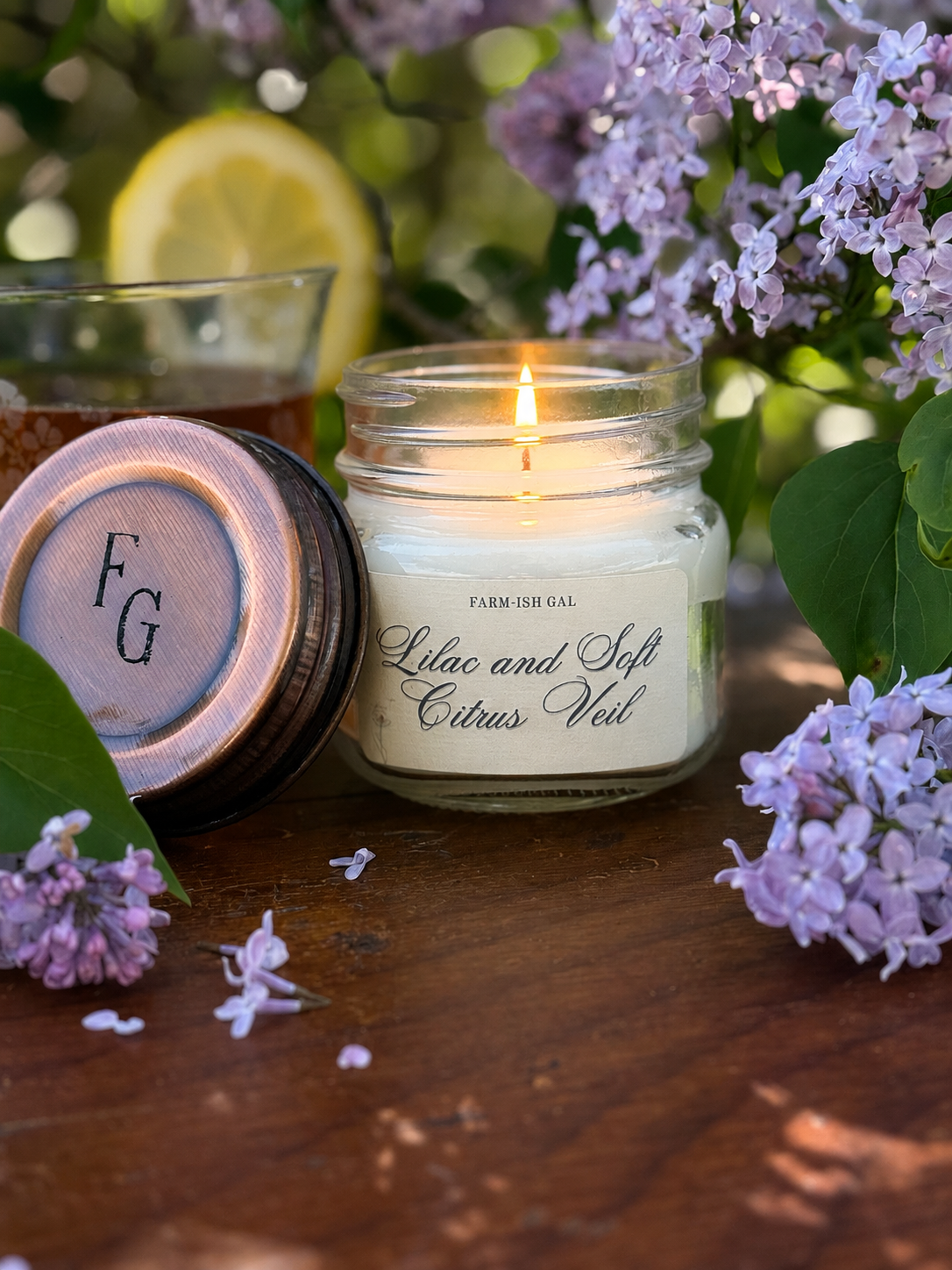 Candle in a jar with a label that says Lilac and Soft Citrus Veil, surrounded by lilacs and a lemon on a wooden surface.