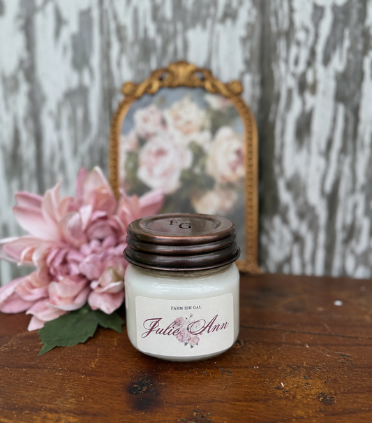 candle jar  labeled 'Julie Ann' with a copper lid on a wooden surface, pink flowers, and a decorative framed picture in the background.