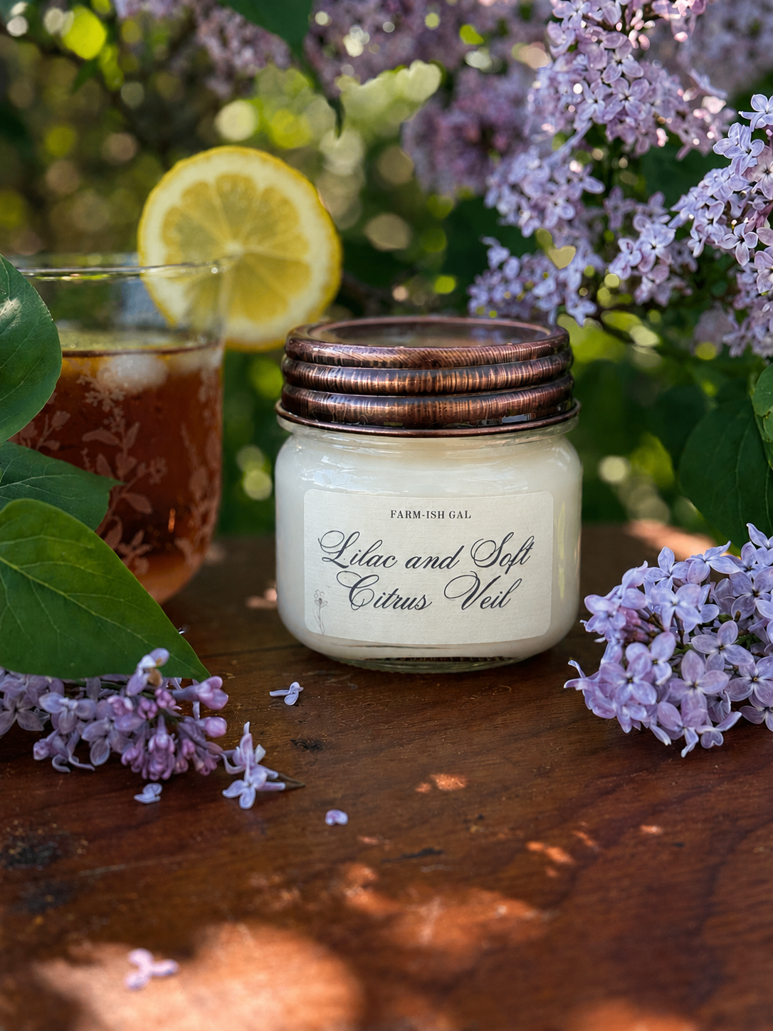 Jar candle with 'Lilac and Soft Citrus Veil' label, surrounded by lilacs and a glass of iced tea with lemon.