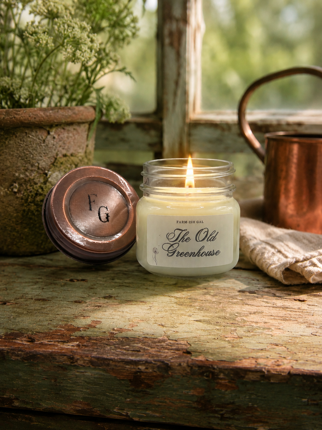 Candle in a jar labeled 'The Old Greenhouse' on a wooden surface with a rustic background.