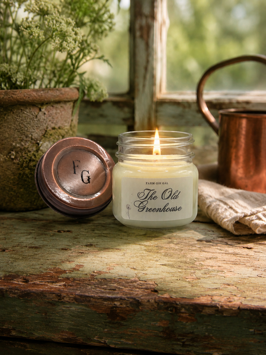 Candle in a jar labeled 'The Old Greenhouse' on a wooden surface with a rustic background.