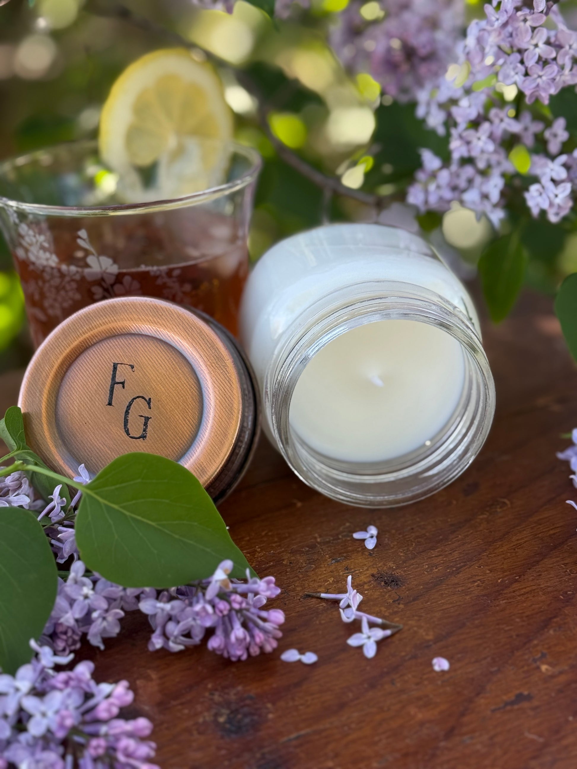 Candle in a glass jar with a copper lid, surrounded by lilacs and a glass of iced tea with a lemon wedge on a wooden surface.
