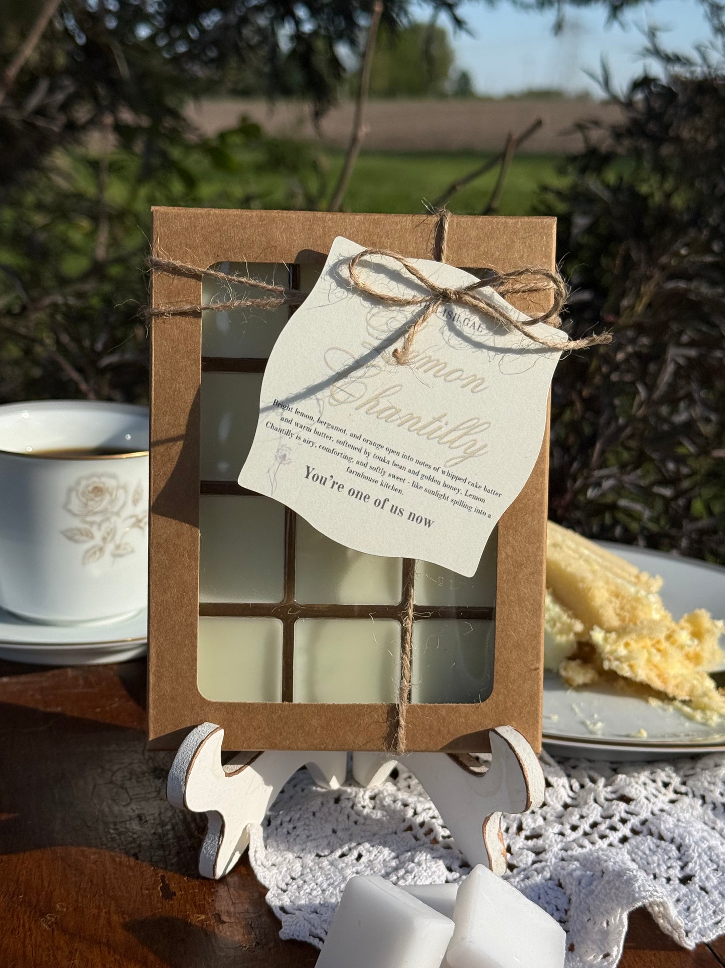 wax melts in a Kraft box with window and tag saying Lemon Chantilly. Cup of coffee, piece of cake and 3 square wax melts on wooden table set around wax melt box.