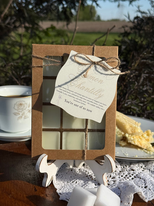 wax melts in a Kraft box with window and tag saying Lemon Chantilly. Cup of coffee, piece of cake and 3 square wax melts on wooden table set around wax melt box.